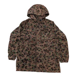 Op de afbeelding: Een camouflagejas met capuchon in bruin, groen en zwart. Het heeft een ritssluiting over de hele lengte, twee borstzakken en een capuchon met trekkoord. De stof heeft een gevlekt patroon, typerend voor kleding in militaire stijl.