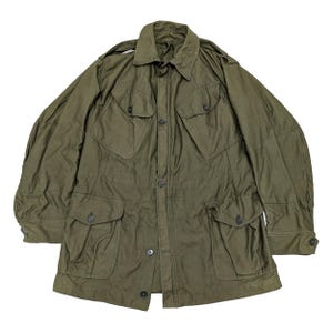 Può includere: Giacca verde oliva in stile militare con multiple tasche frontali e chiusure con bottoni. La giacca presenta un colletto e spalline. Il tessuto sembra essere un materiale resistente, forse cotone.