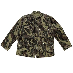 May include: A camouflage jacket in shades of green and brown. The jacket features multiple front pockets with button closures, a button-up front, and a collared neckline. The jacket has long sleeves and a military-inspired design.