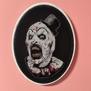May include: A black and white cross-stitch embroidery of a creepy clown with a white face, red lips, and blood splatters. The clown is wearing a black top hat and a white collar.