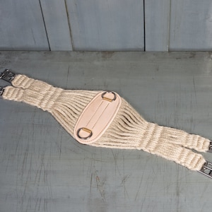May include: A beige leather and rope girth for a horse. The girth has two metal buckles and a leather strap with two metal rings.