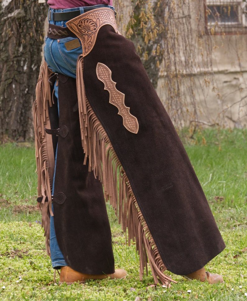 Pu&ograve; includere: Un paio di chaps in pelle scamosciata marrone con dettagli in frangia e un design in pelle traforata. Le chaps sono indossate sopra un paio di jeans blu e una cintura marrone.