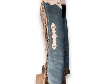 Op de afbeelding: Een paar blauwe suède chaps met franjes en een tan lederen rand. De chaps hebben een zilveren knoopdetail en een riemlus.