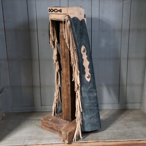 May include: A pair of blue suede chaps with fringe and brown leather accents. The chaps are displayed on a wooden stand.