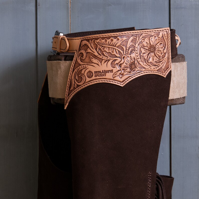 Pu&ograve; includere: Chaps da equitazione in pelle marrone con un design floreale decorativo e il testo "Sedlarstvi Spurny" sul davanti.