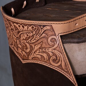 Pu&ograve; includere: Un paio di chaps in pelle marrone con una cintura in pelle beige e una fibbia argentata. I chaps hanno un design floreale inciso nella pelle.
