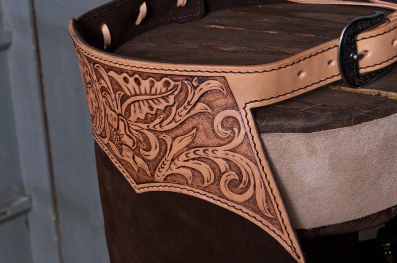 Pu&ograve; includere: Un paio di chaps in pelle marrone con una cintura in pelle beige e una fibbia argentata. I chaps hanno un design floreale inciso nella pelle.