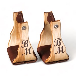 May include: A pair of wooden stirrup hangers with brown leather accents. The hangers are engraved with the initials "BM".
