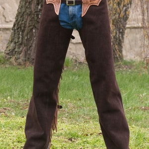 Pu&ograve; includere: Chaps in pelle marrone con frange e un design lavorato. I chaps hanno un passante per cintura e una chiusura a fibbia. I chaps sono indossati sopra un jeans blu.