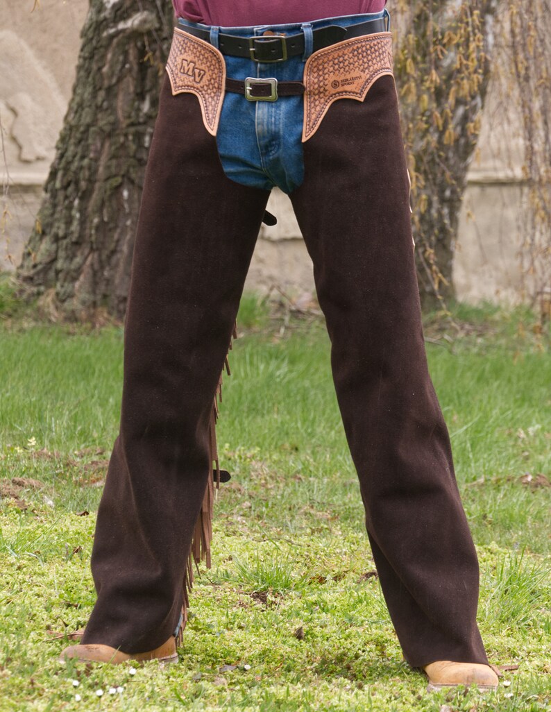 Pu&ograve; includere: Chaps in pelle marrone con frange e un design lavorato. I chaps hanno un passante per cintura e una chiusura a fibbia. I chaps sono indossati sopra un jeans blu.