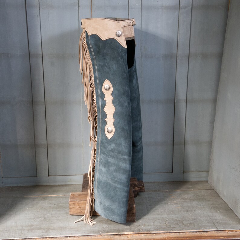 May include: A pair of blue suede chaps with fringe and leather accents. The chaps have a tan leather waistband and are decorated with silver conchos.
