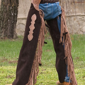 Pu&ograve; includere: Chaps in pelle marrone con frange e cuciture decorative. I chaps sono indossati sopra un paio di jeans blu.