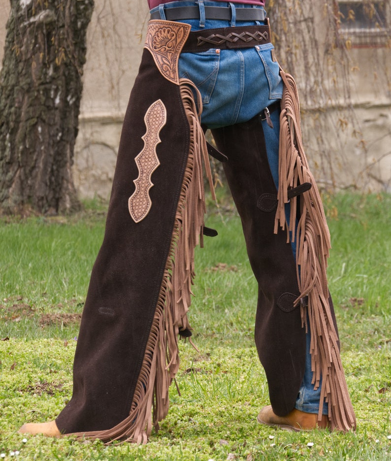 Pu&ograve; includere: Chaps in pelle marrone con frange e cuciture decorative. I chaps sono indossati sopra un paio di jeans blu.