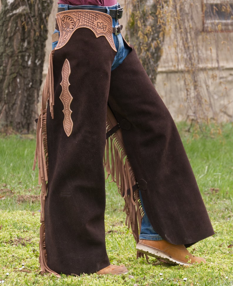 Pu&ograve; includere: Chaps in pelle marrone con dettagli a frangia e un design lavorato. Le chaps hanno una chiusura a fibbia e vengono indossate sopra i jeans.