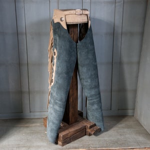 May include: A pair of blue suede chaps with tan fringe and a leather belt. The chaps are displayed on a wooden stand.
