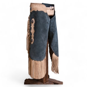 Può includere: Un paio di chaps in pelle scamosciata blu scuro con frange color sabbia e dettagli in pelle. I chaps hanno un passante per la cintura e una chiusura a fibbia.