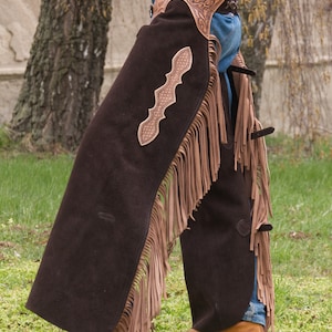 Pu&ograve; includere: Un paio di chaps in pelle marrone con frange e un design in pelle traforata. I chaps sono indossati sopra un jeans blu e stivali marroni.