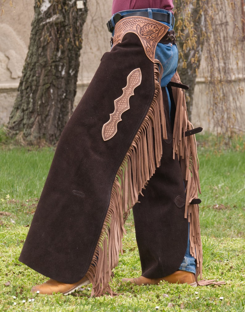 Pu&ograve; includere: Un paio di chaps in pelle marrone con frange e un design in pelle traforata. I chaps sono indossati sopra un jeans blu e stivali marroni.