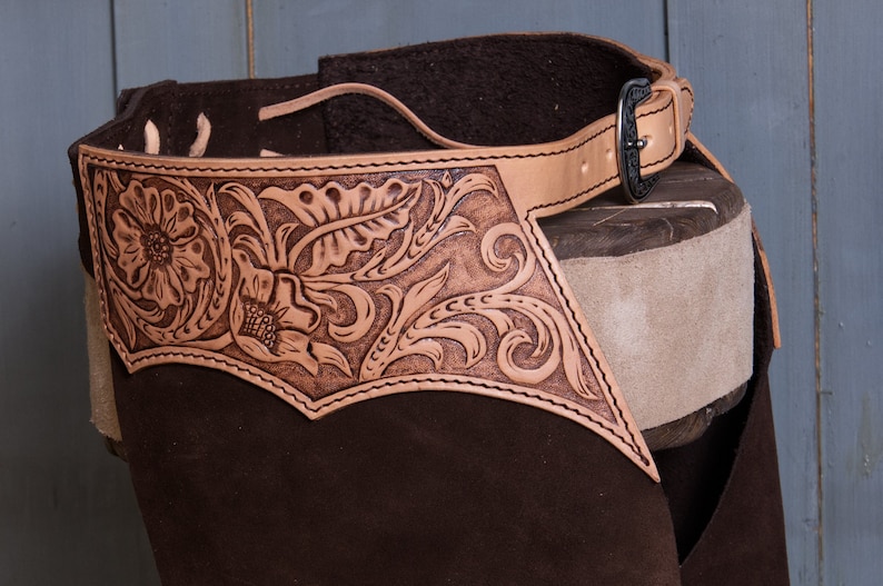 Pu&ograve; includere: Chaps in pelle marrone con un design decorativo a rilievo che presenta motivi floreali e a volute. I chaps hanno una cintura con una fibbia argentata.
