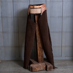 Pu&ograve; includere: Chaps in pelle marrone con frange e una cintura in pelle lavorata. I chaps sono esposti su un supporto in legno.