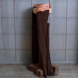 Pu&ograve; includere: Chaps in pelle marrone con frange e una cintura in pelle lavorata. I chaps sono appesi a un supporto in legno su uno sfondo grigio.