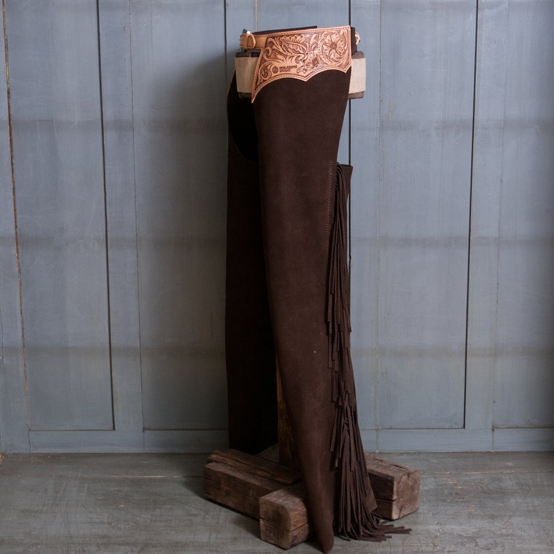 Pu&ograve; includere: Chaps in pelle marrone con frange e una cintura in pelle lavorata. I chaps sono appesi a un supporto in legno su uno sfondo grigio.