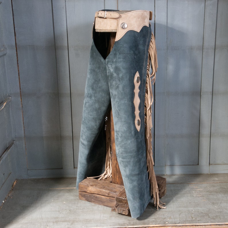 May include: A pair of blue suede chaps with fringe and brown leather accents. The chaps are displayed on a wooden stand.