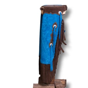 Può includere: Un paio di chaps in pelle scamosciata blu con frange marroni e dettagli argentati. I chaps sono esposti su un supporto in legno.