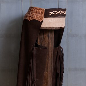 Pu&ograve; includere: Chaps in pelle marrone con frange e un design in pelle traforata. I chaps sono appesi a un palo di legno.