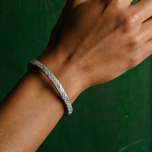 Op de afbeelding: Een zilveren armband met een getextureerd, geweven ontwerp. De armband zit om een pols.
