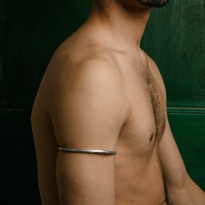 Op de afbeelding: Een man zonder shirt met een baard en een neusring poseert voor een groene houten deur. Hij draagt een zilveren armband op zijn linkerarm.