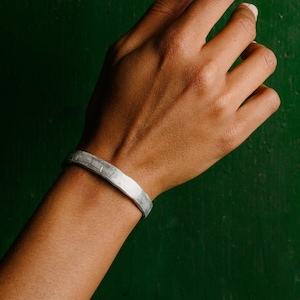 Op de afbeelding: Een zilveren armband met een gehamerde textuur op de pols van een persoon tegen een groene achtergrond.