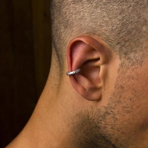 Könnte beinhalten: Eine silberfarbene Ohrmanschette mit einer texturierten, gehämmerten Oberfläche. Der kreisförmige Metallring weist ein unregelmäßiges, organisches Finish auf und ist so gestaltet, dass er sicher am äußeren Knorpel des Ohrs sitzt, ohne ein Piercing.