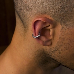 Könnte beinhalten: Ein glatter, kreisförmiger silberfarbener Metall-Creolen-Ohrring, der im oberen Knorpelbereich des Ohrs getragen wird. Der Ohrring hat ein schlichtes, durchgehendes Ringdesign mit polierter Oberfläche und sitzt fest in der äußeren Ohrfalte.