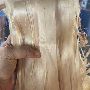 Puede incluir: Un manojo de tiras de material natural de color beige claro, fibrosas y translúcidas. Las tiras son largas, planas y tienen una textura suave y similar al papel con bordes deshilachados y delicados. El material está apilado en capas.