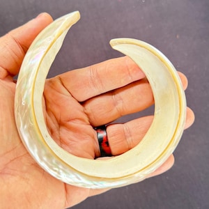 Puede incluir: Una pulsera nacarada, abierta, con un diseño curvo. La pulsera es de color blanco crema con reflejos iridiscentes. El diseño abierto permite llevarla fácilmente. La pulsera tiene un diámetro de aproximadamente 6,35 cm.