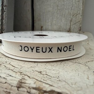 May include: A spool of off-white fabric ribbon featuring the black printed text JOYEUX NOEL. The ribbon is wound around a white circular core, displaying a woven texture and clear, bold lettering centered along the length of the material.