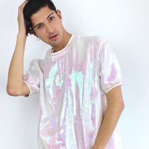 May include: A white iridescent sequined short-sleeved shirt with a pink undertone. The shirt has a round neckline and a white trim around the neckline.