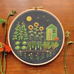May include: A circular embroidery hoop containing a black fabric panel with a hand-stitched garden scene. The design features a green shed, sunflowers, vegetable patches with leafy greens, climbing vines, and a yellow sun against a dark background.