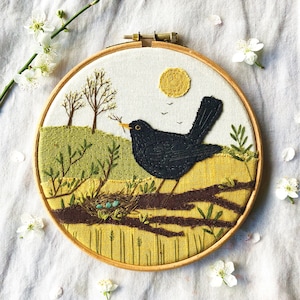 Peut inclure: Art de broderie en forme de cercle représentant un merle perché sur une branche avec un nid d'œufs bleus. L'arrière-plan est un paysage jaune et vert avec un soleil et des arbres.