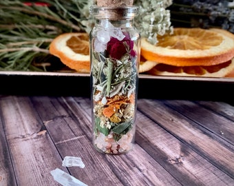May include: A clear glass bottle with a cork stopper filled with dried herbs, flowers, and crystals. The bottle is sitting on a wooden surface.