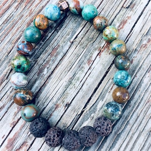 May include: A beaded bracelet featuring a variety of colorful, round stones. The bracelet includes blue, green, brown, and patterned beads, along with dark, porous lava beads. The bracelet is displayed on a weathered wooden surface.