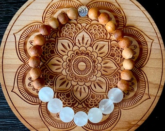Peut inclure: Un bracelet en bois avec un charme argenté et des perles blanches. Le bracelet est sur un fond en bois avec un motif de mandala.