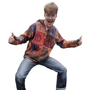 May include: A man wearing a multi-coloured patchwork shirt with a hood, blue jeans, and white trainers with black accents. He is smiling and giving a thumbs up with both hands.