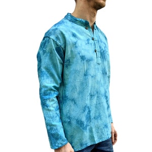 May include: A long-sleeved, tie-dye shirt in shades of turquoise and blue. The shirt has a mandarin collar, a button-down placket, and a small pocket on the chest. The model is wearing blue jeans.