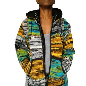 May include: A colorful, hooded zip-up jacket with a patchwork design. The jacket features a mix of yellow, teal, gray, and black knitted sections. It has a black zipper, hood lining, and pocket trim. The jacket is worn over a gray tank top and blue jeans.