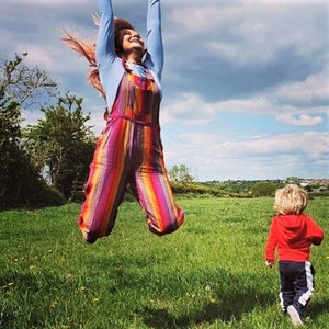 May include: A woman in a rainbow striped jumpsuit jumps in a field with a young boy in a red hoodie and black pants walking towards her.