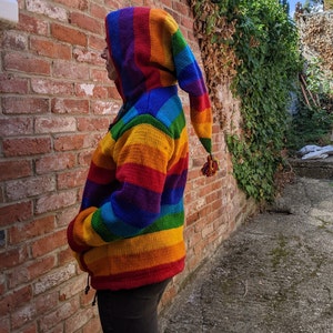 May include: A rainbow striped knitted hoodie with a long pointed hood. The hoodie has a zip fastening and two pockets.