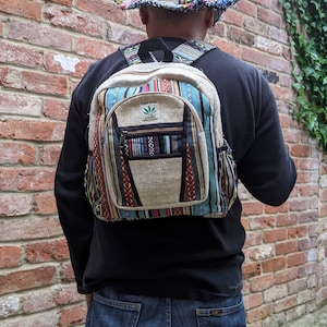 May include: A person wearing a black shirt and blue jeans is wearing a hemp backpack with a multi-colored striped design. The backpack has a small pocket on the front with the text "PURE HEMP THE REAL DEAL MADE IN INDIA". The person is also wearing a multi-colored hemp hat.
