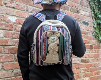 May include: A person wearing a black shirt and blue jeans is wearing a hemp backpack with a multi-coloured striped design. The backpack has a small round pocket on the front with the words "Pure Hemp" and a green marijuana leaf logo.